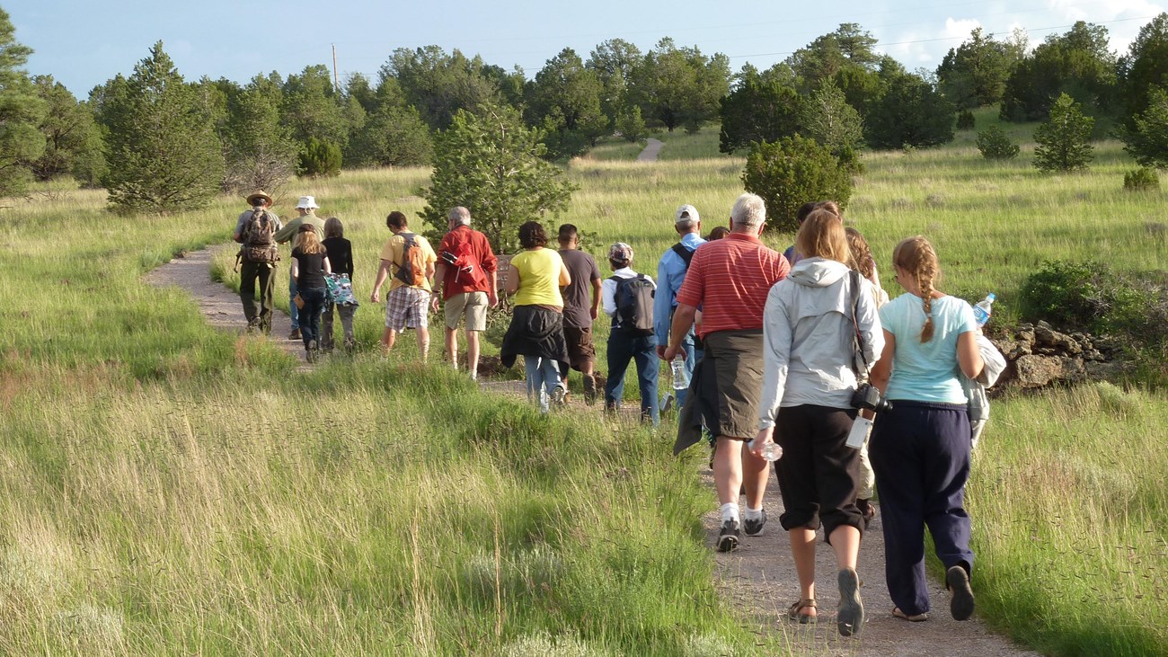 A group of hikers heads down a gravel trail through grass with trees in the distance