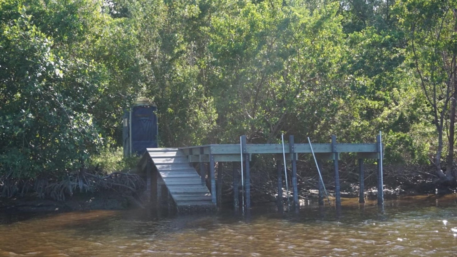 Murky brown water runs up against a mangrove shoreline, with a pitched dock and elevated platform