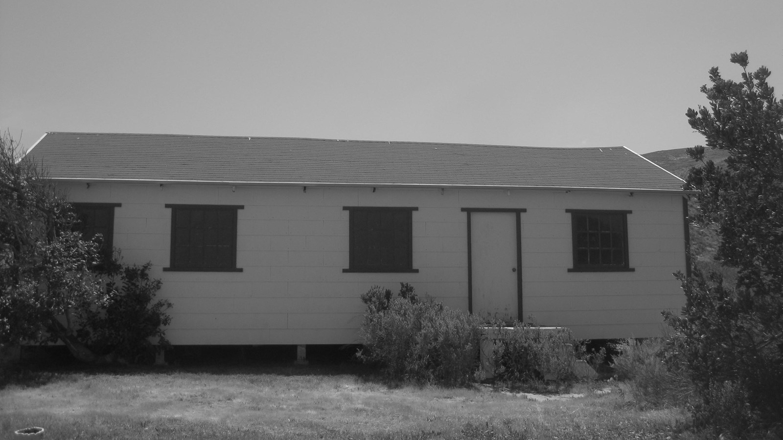 long wood building with 4 windows and door with bushes on each side.