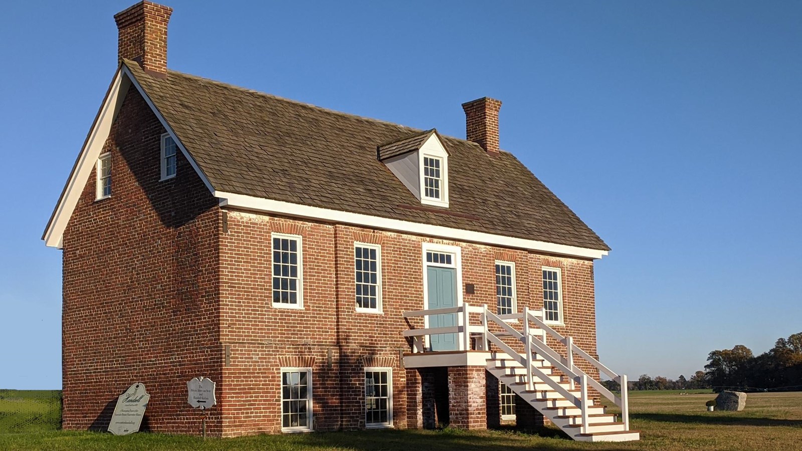 A brick house in a grass field with a steep roof, white windows, and a blue door accessed by stairs.