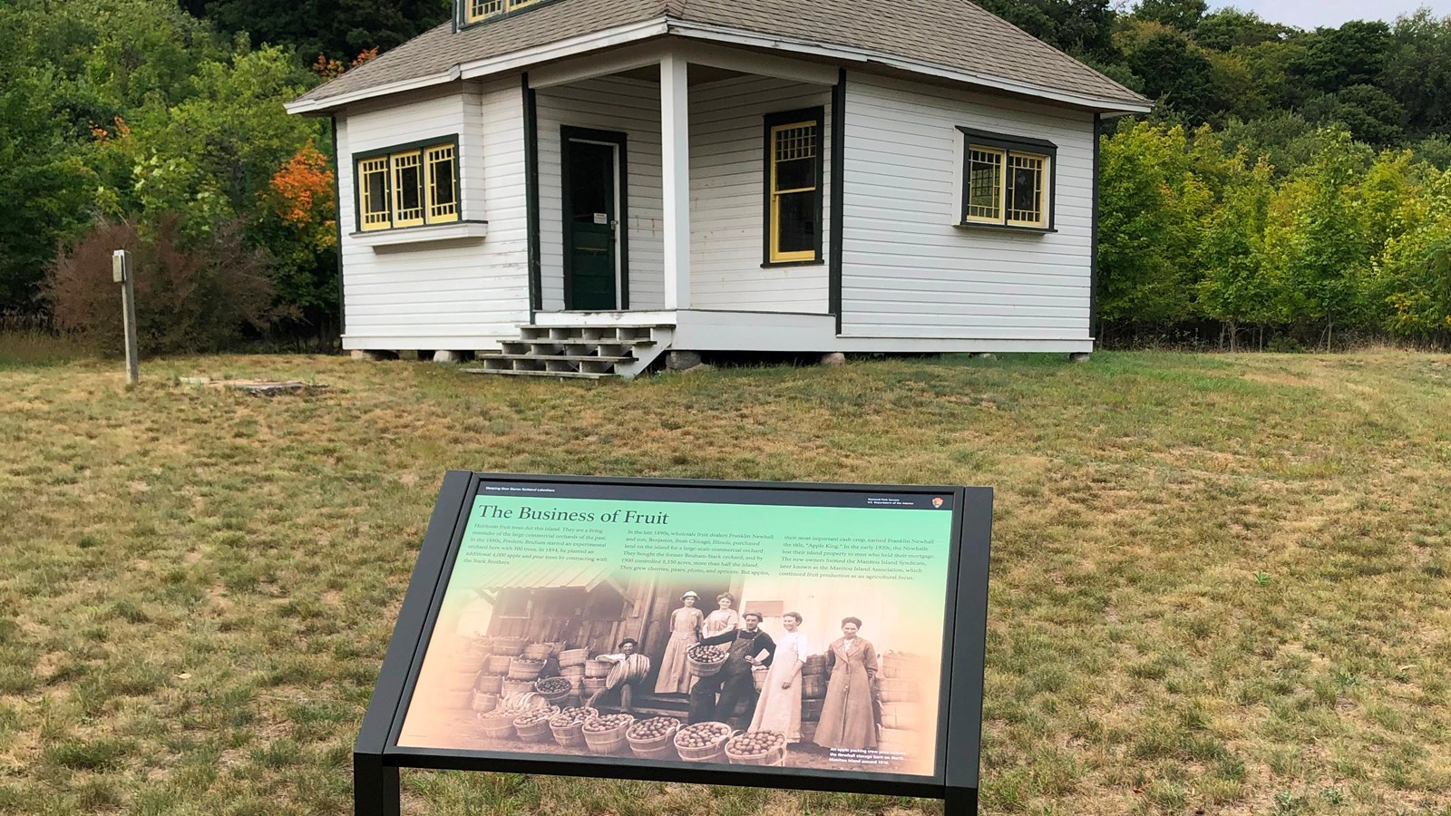 An informational sign stands in front of a small white historic building with a brown roof