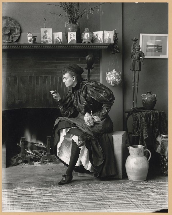Full-length portrait, seated in front of fireplace, holding a cigarette and a beer stein