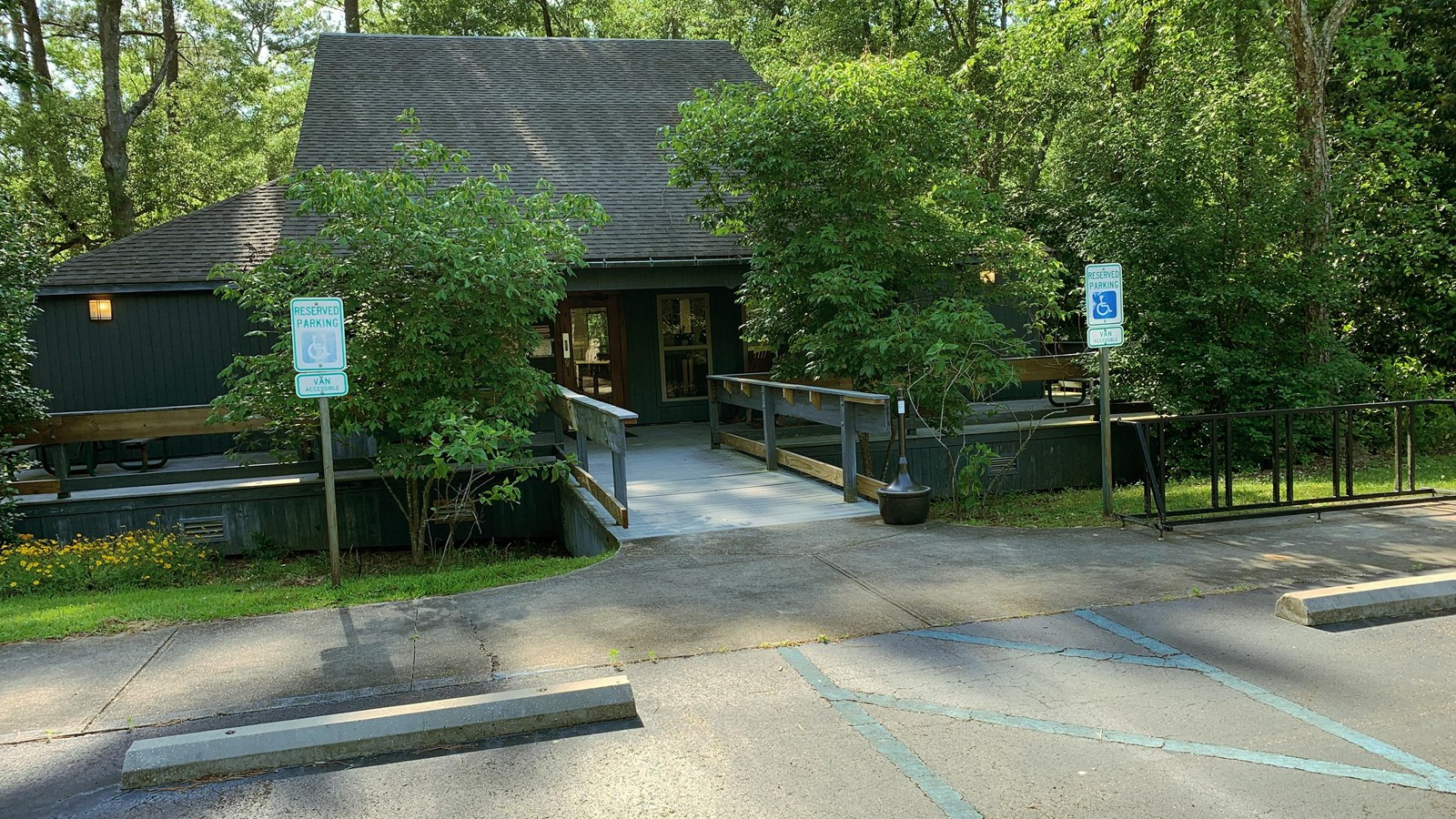 a parking lot leads to a railed entrance to a small building surrounded by green trees.