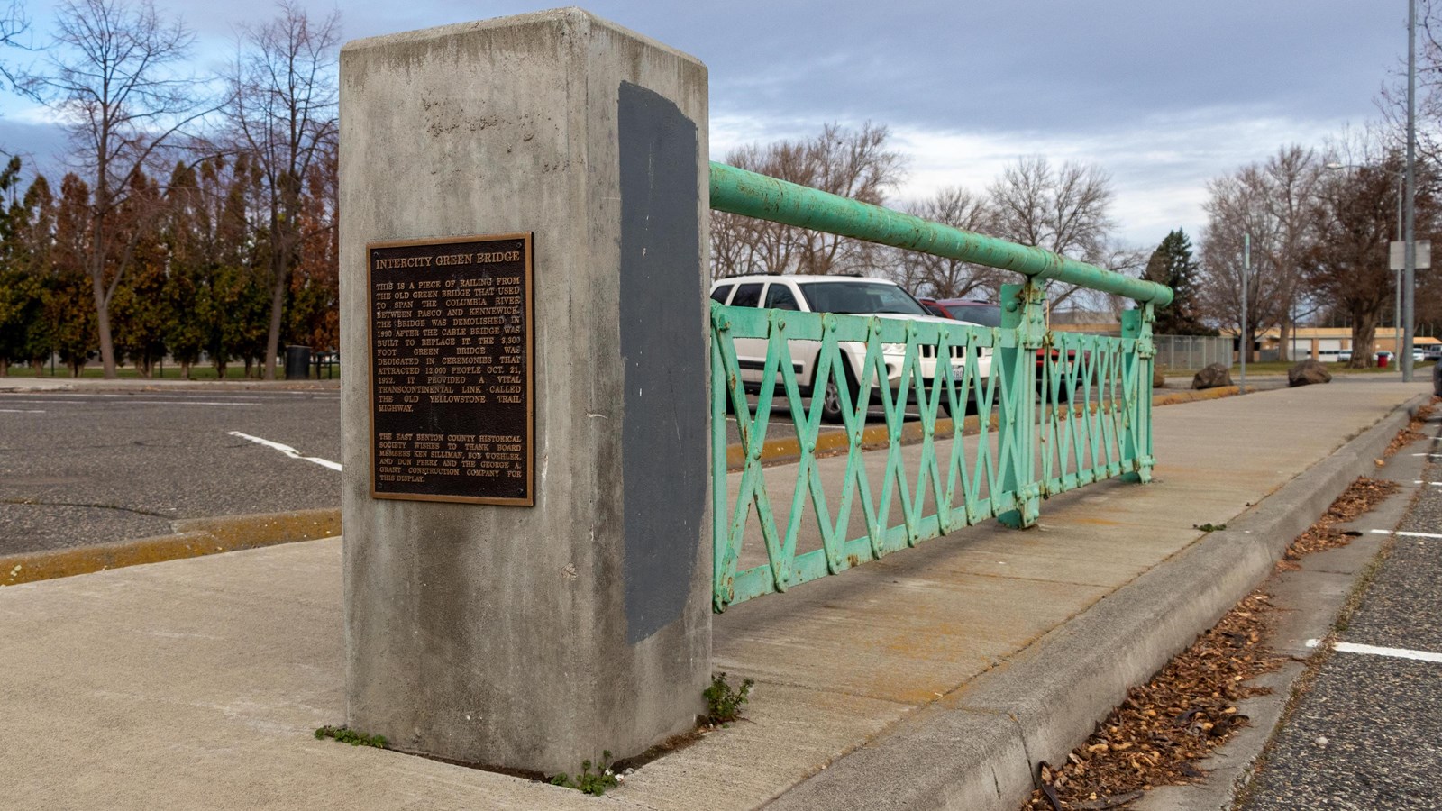 A green railing of a bridge is attached to a cement pillar with a plaque describing the bridge.  