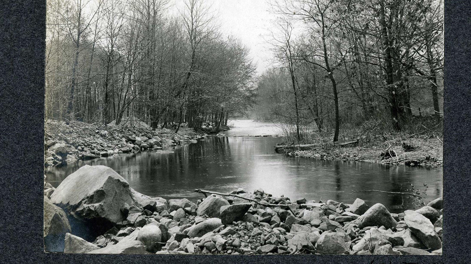 Black and white of river lined with rocks and trees.
