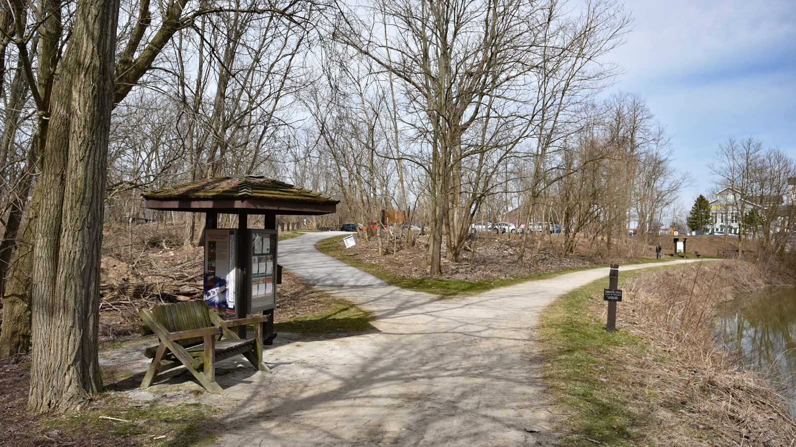 A three-sided kiosk and bench are at a fork. The trail on the right is beside a water-filled canal. 