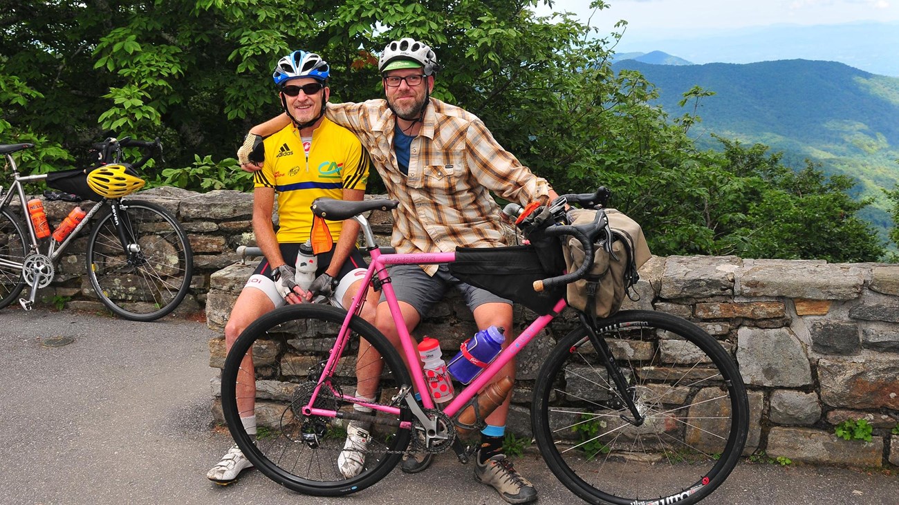 Two bicyclists sit on a stone wall near their bikes. Forest and mountains in background.