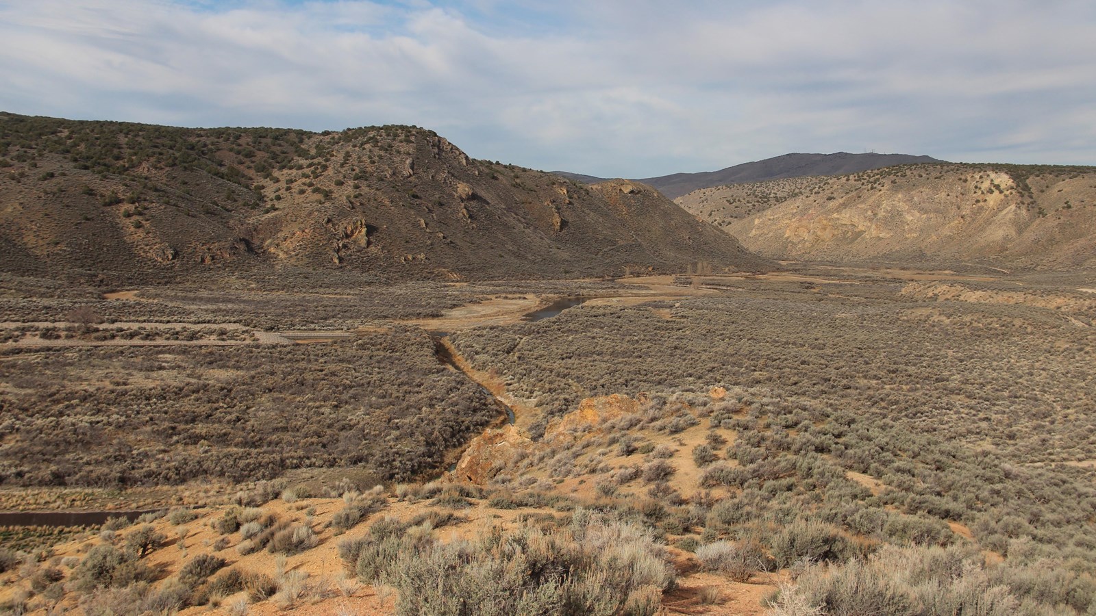 A narrow channel winds through a sagebrush plain toward steep bluffs in the distance.