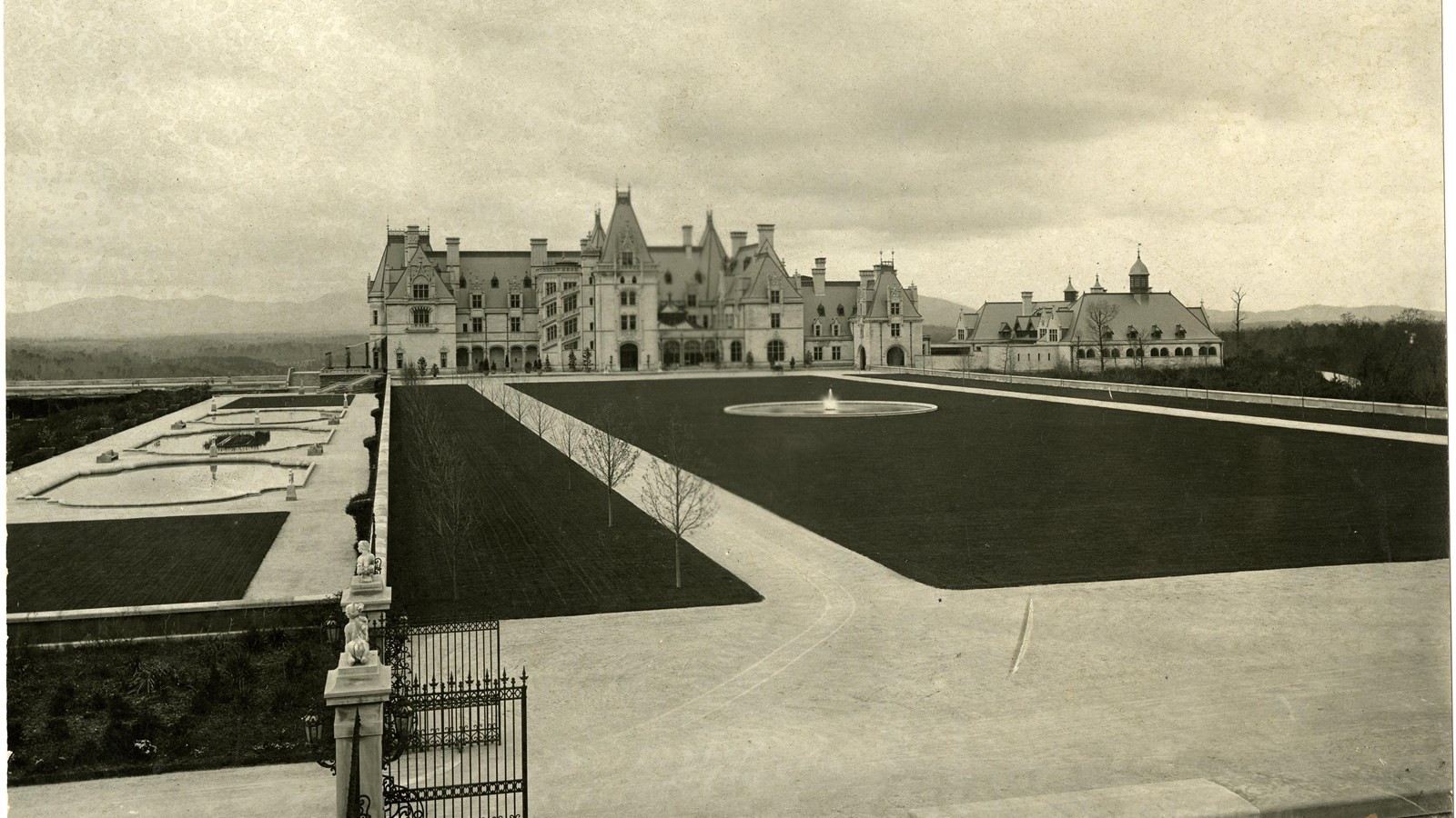 Black and white of large home in front of large flat grassy area with straight paths on side. 