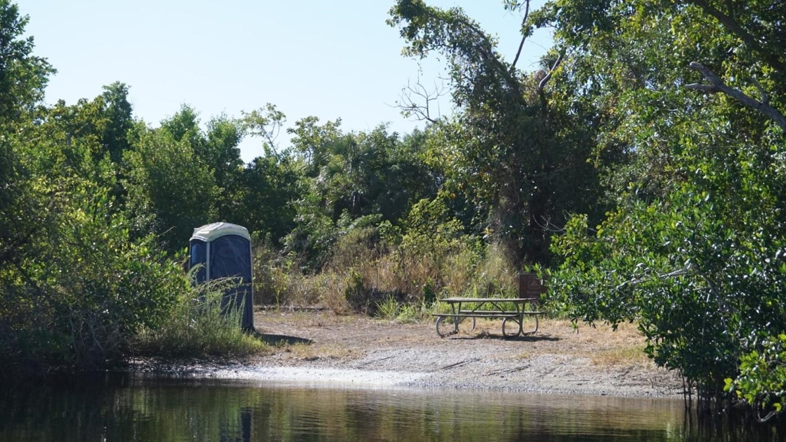 A small beach clearing surrounded by shrubby green vegetation. A blue vault toilet is observed