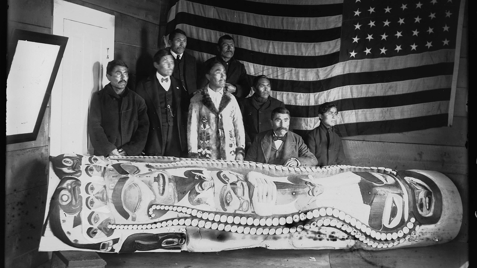 A historic photo eight men and a US flag arranged behind Sleeping Man Pole. The pole is horizontal.