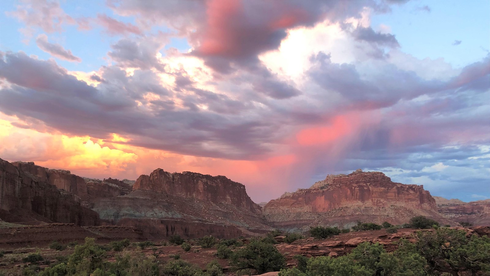 Juniper trees dot the foreground, with distant red-brown cliffs and colorful cloudy sky at sunset.