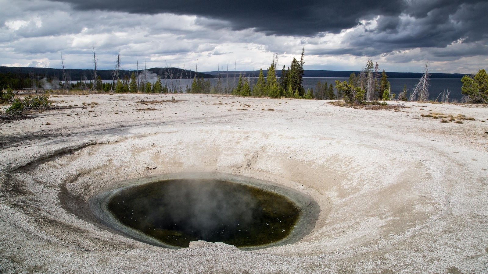 A deeply set and darkly colored hot spring sits in a geyser basin near a large lake.