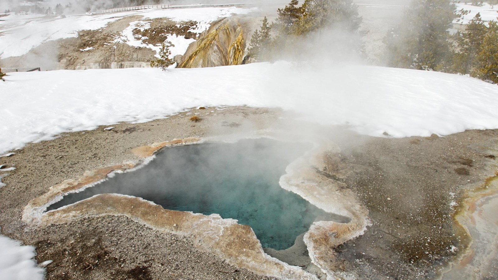 An irregular shaped, turquoise blue hot spring sits in a thermal basin covered in patchy snow.