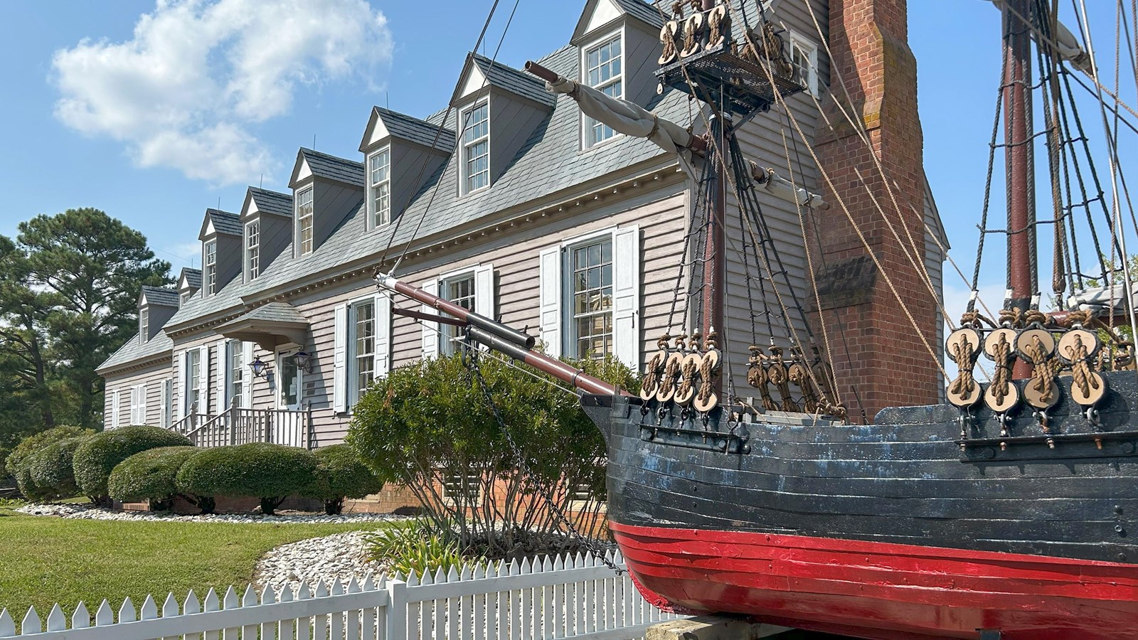 A ship model stands before a gray historic building, bushes, and a picket fence.