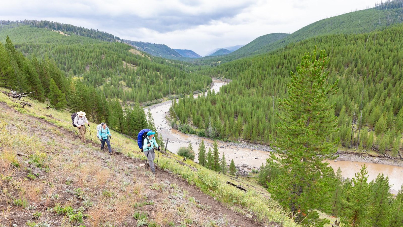 Three backpackers walk along a trail above a forested river valley.
