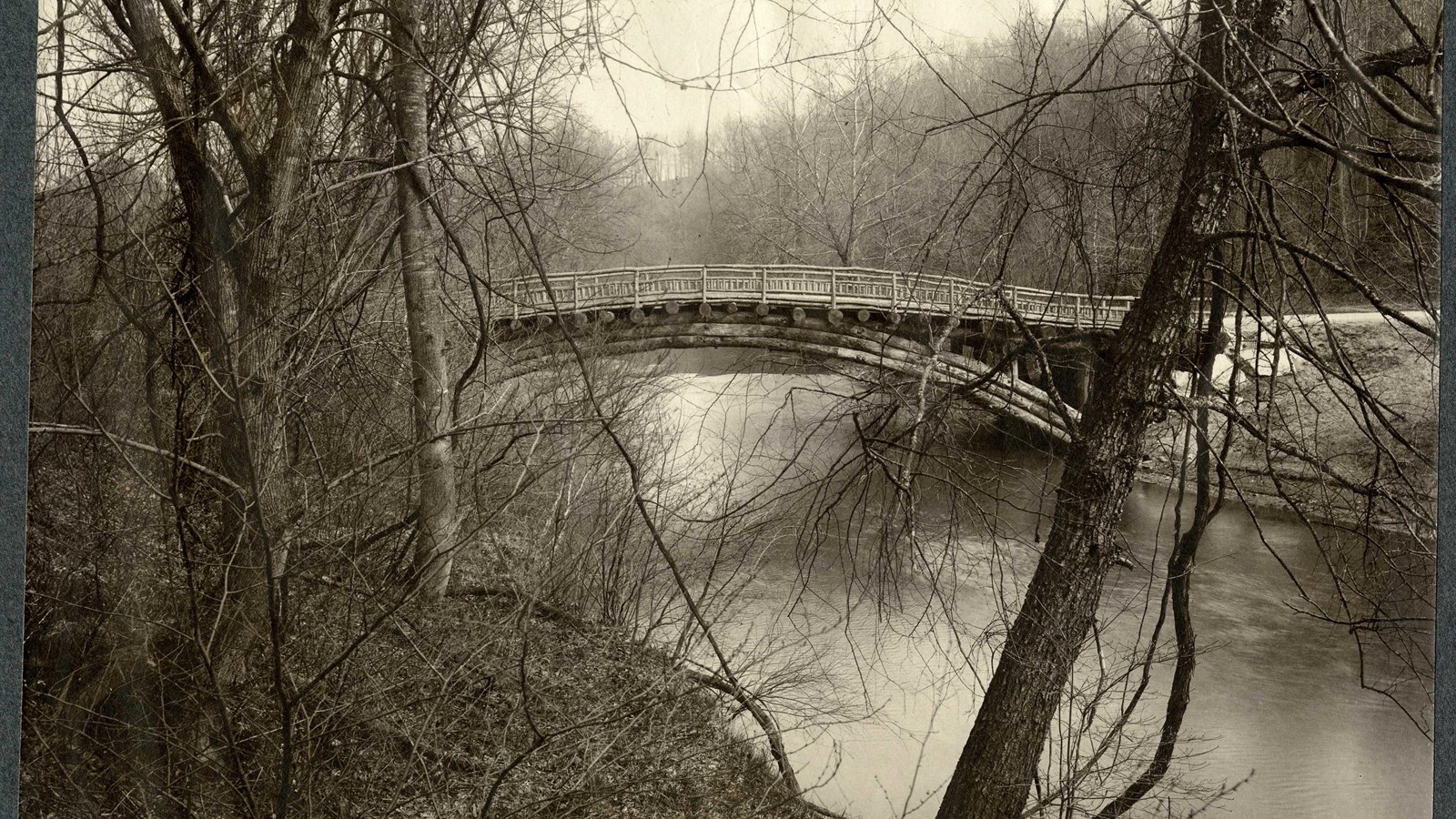 Black and white of bridge over water with trees on one side, hilly going down to water on other