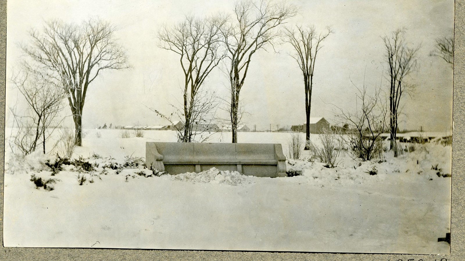 Black and white of stone bench on snowy flat area with some trees behind it with no leaves. 