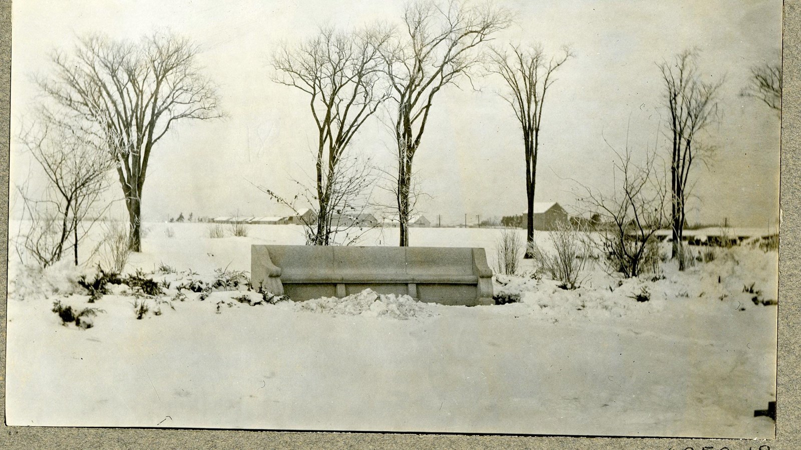 Black and white of stone bench on snowy flat area with some trees behind it with no leaves.