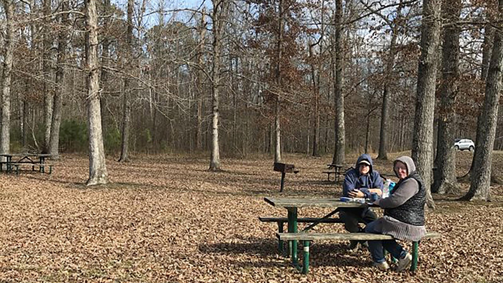A picnic table with two people bundled in hats and coats eating, in a the midst of leafless trees.