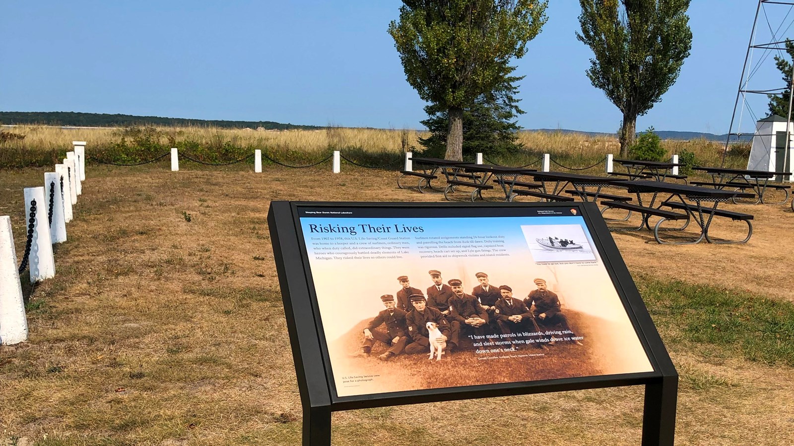 An informational sign stands in front of a fence-lined grassy field with two trees in the background