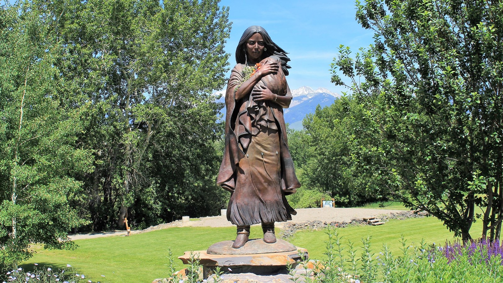 A bronze statue of Sacajawea holding her child amid green grass and trees