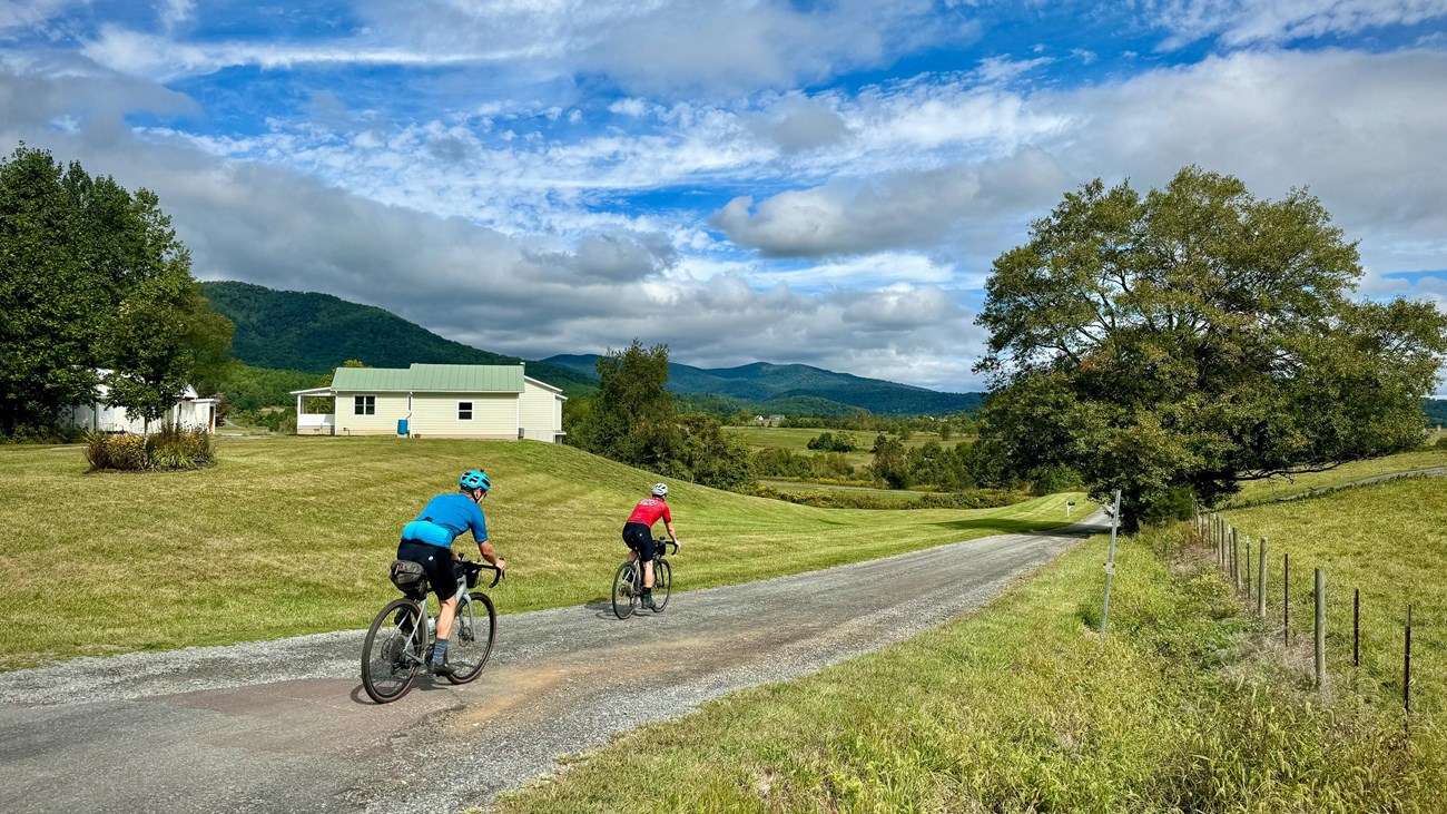 Two cyclists on a gravel road with hills and a house in the background under a cloudy blue sky.