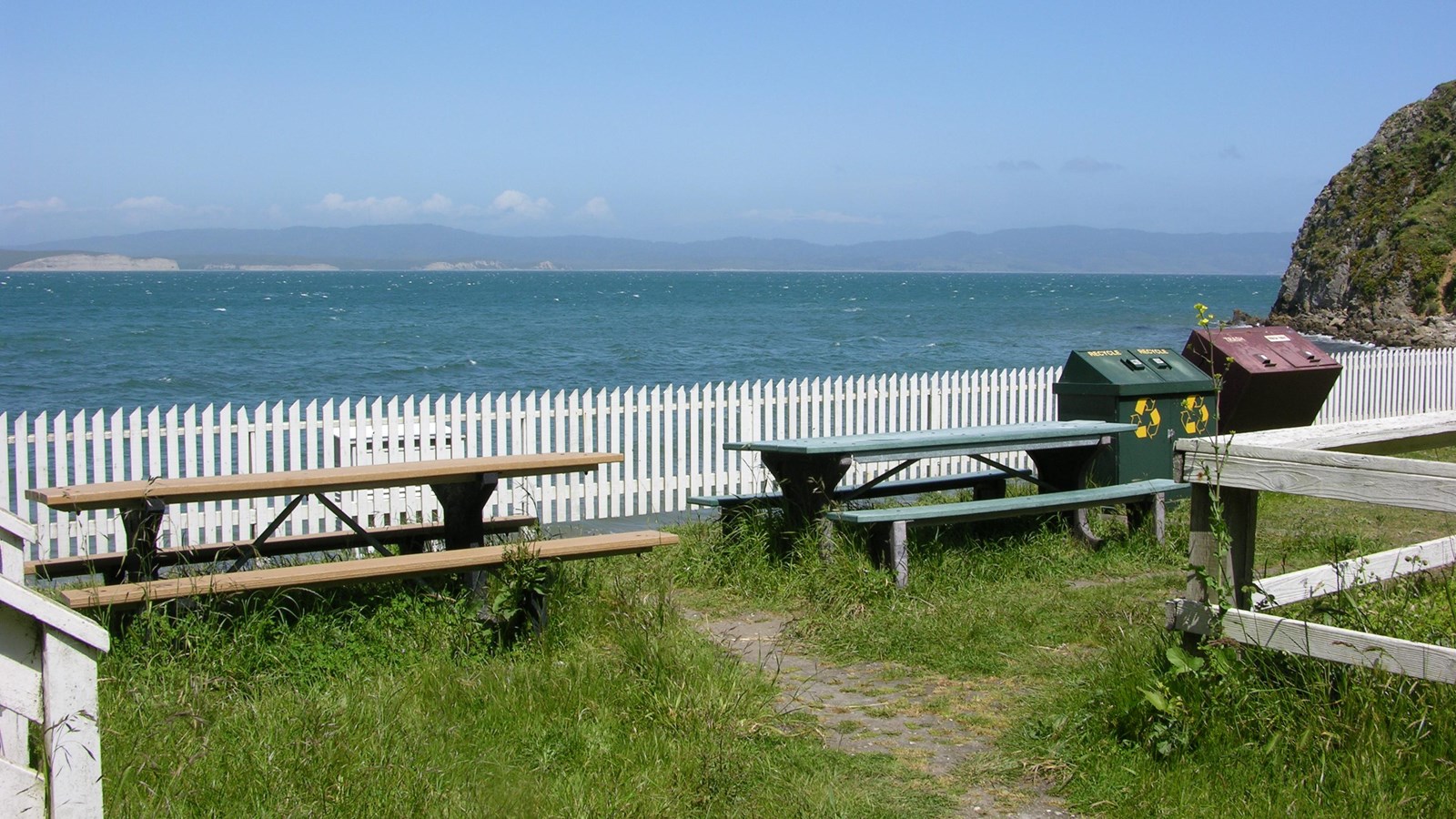 Two picnic tables next to refuse containers and a white picket fence with a bay in the background.
