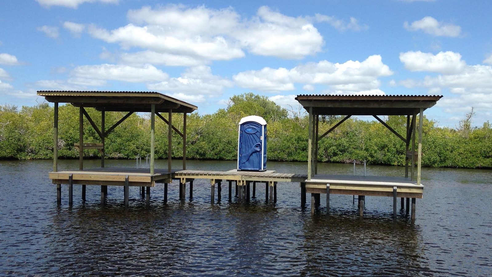 A open air platform elevated over the water with two roofs and a blue portable toilet in between