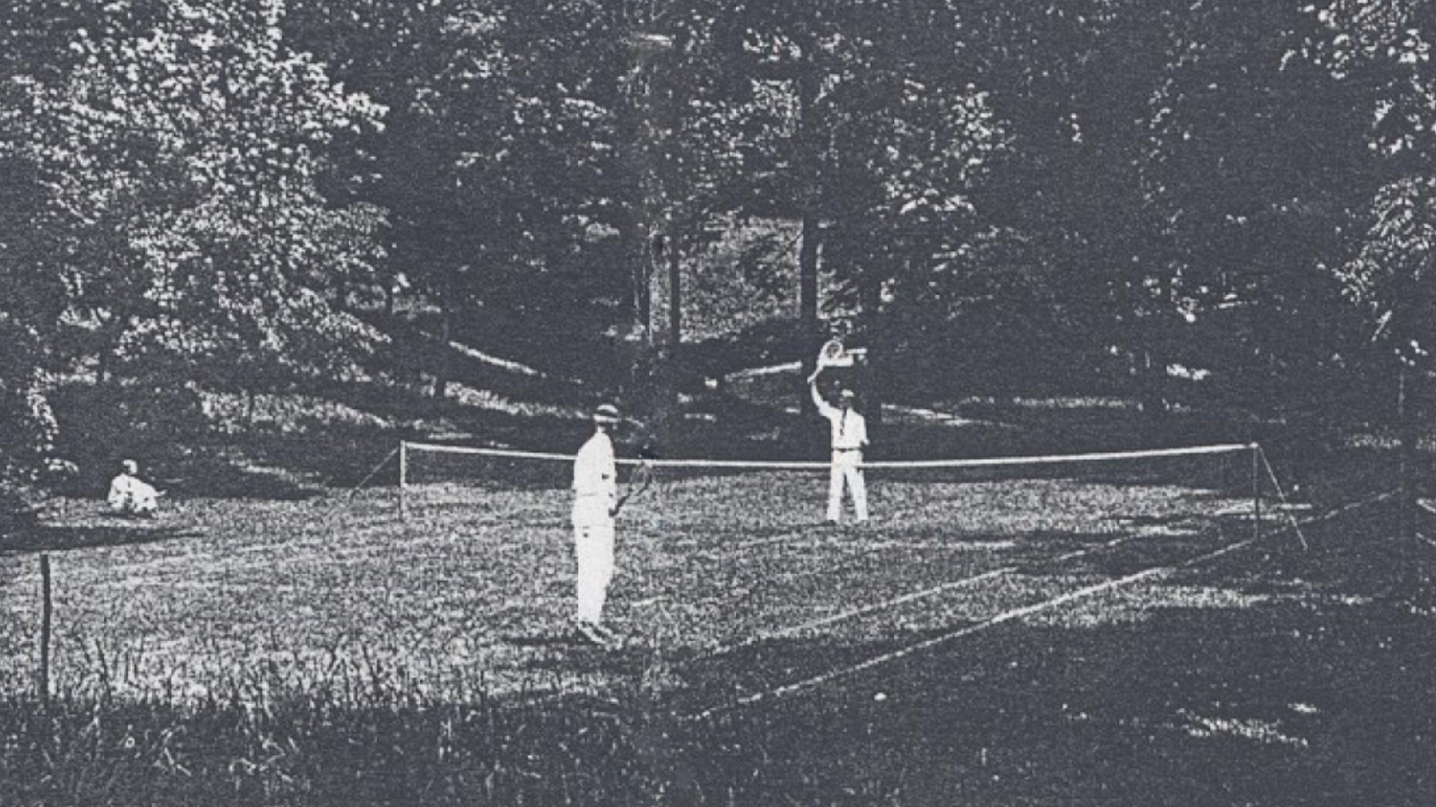 A black and white photo of two men in white clothes on a tennis court surrounded by woods.