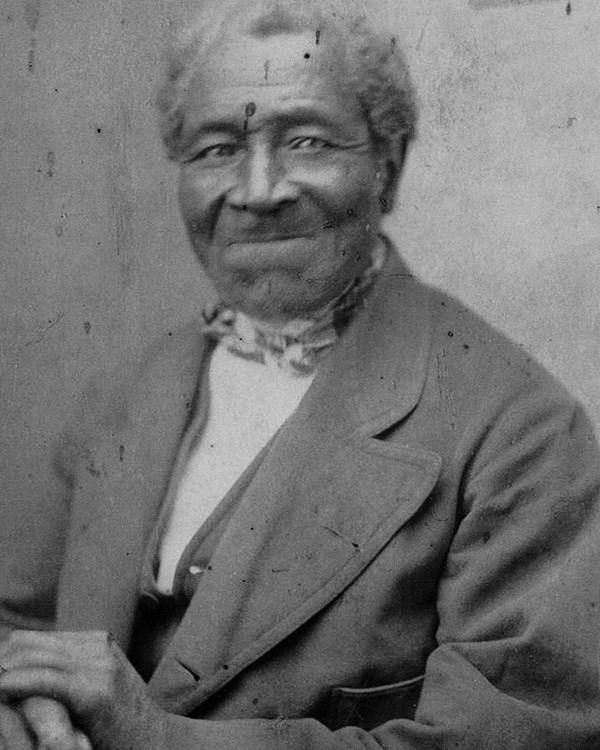 Older African American male in 19th century jacket, seated holding a cane.