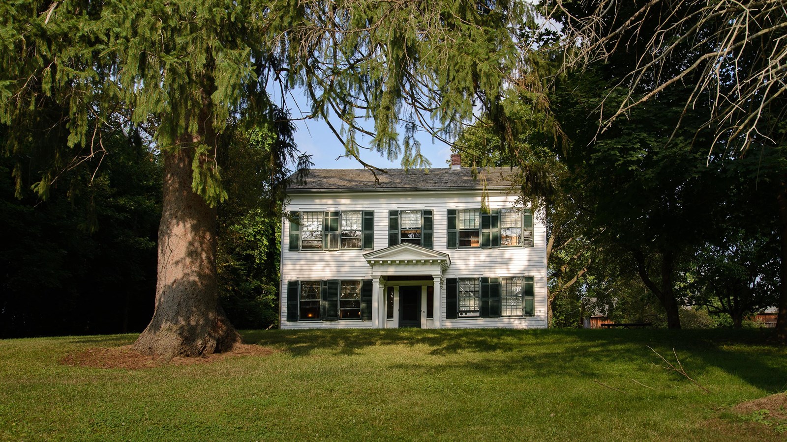 A historic white house with green shutters and door sits atop a low hill, surrounded by tall trees.