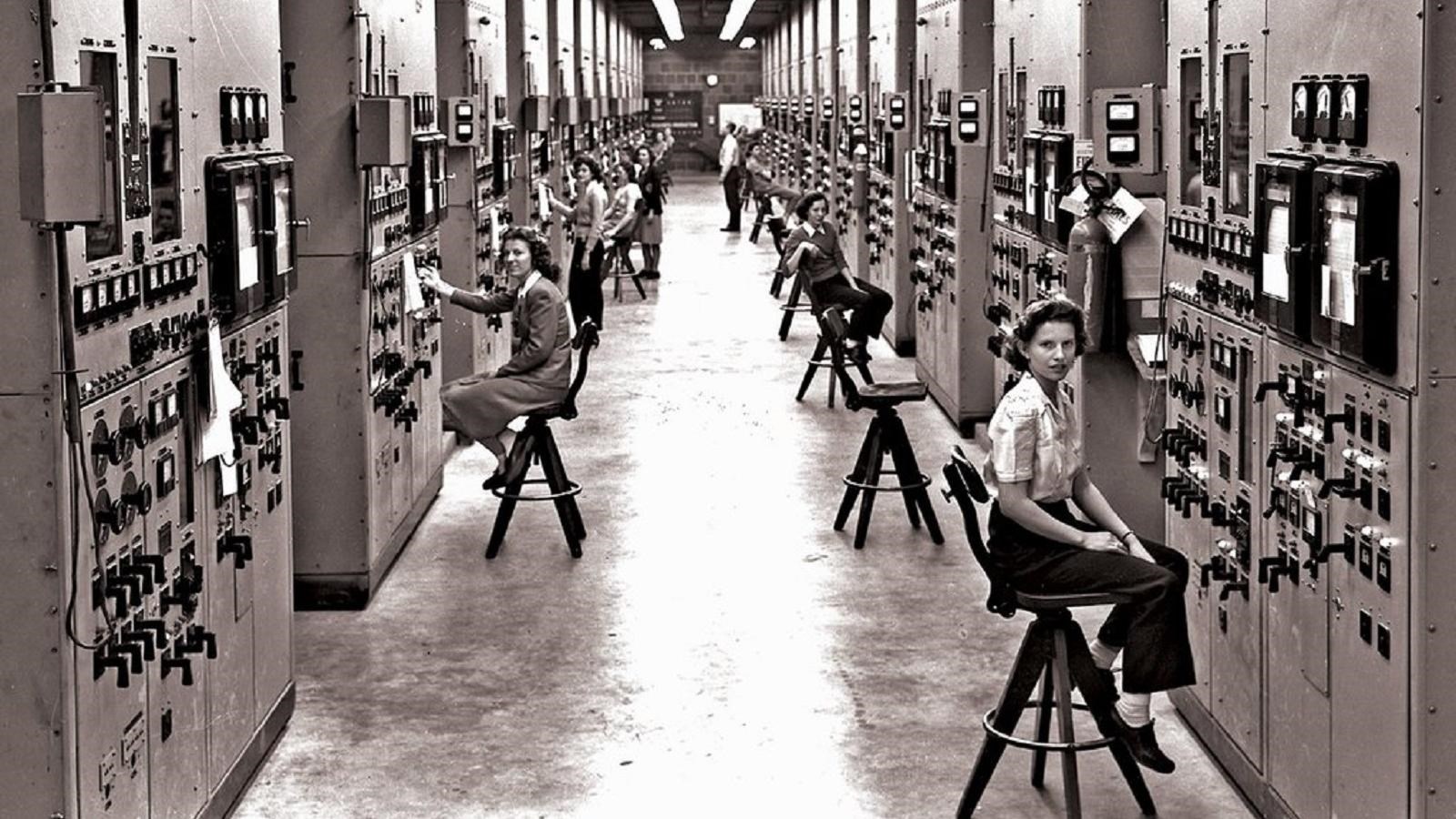 women sitting on stools in a large room working on very large mainframe computers