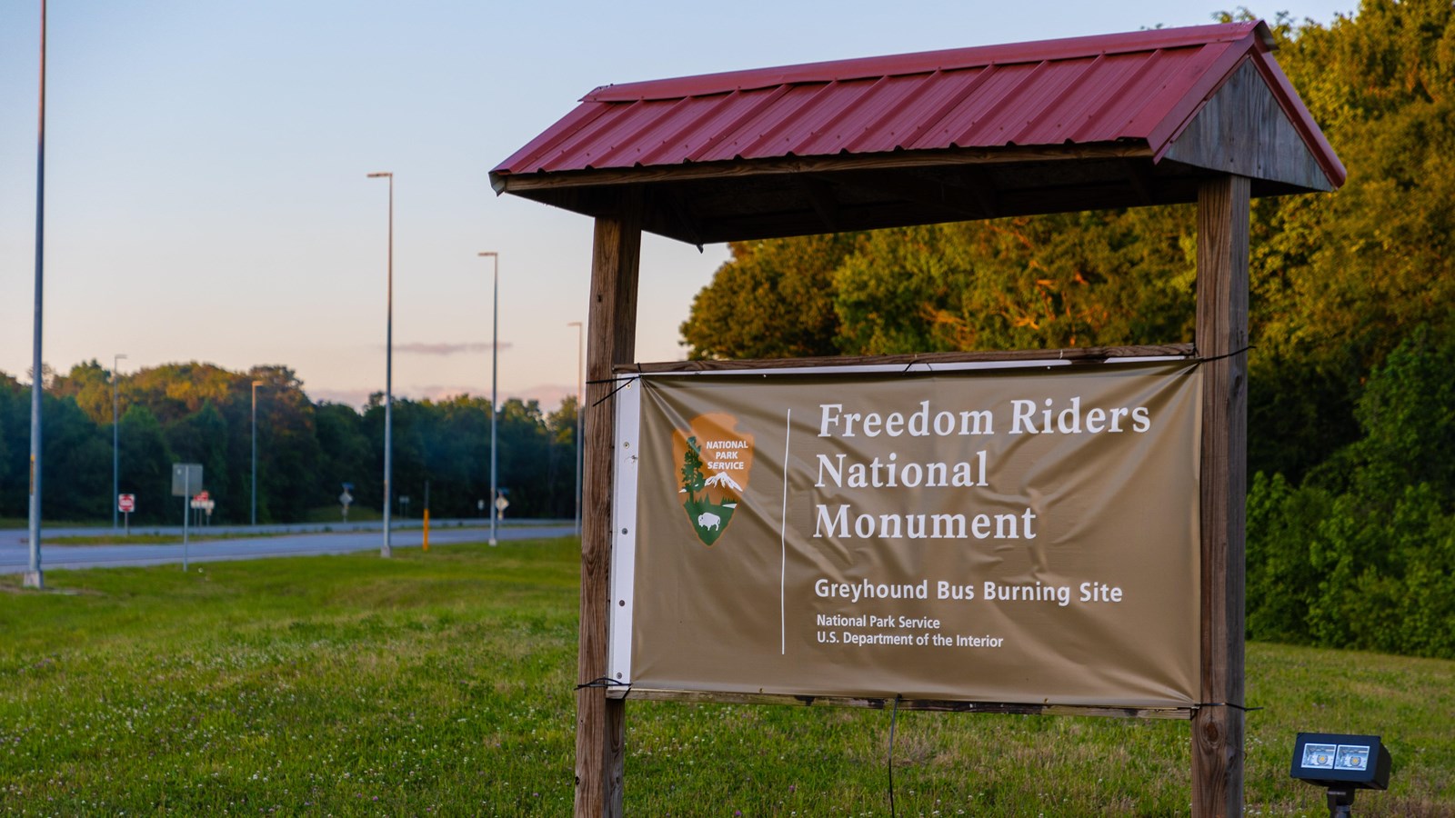 A brown NPS sign, under a red metal awning, on a green field near a highway.