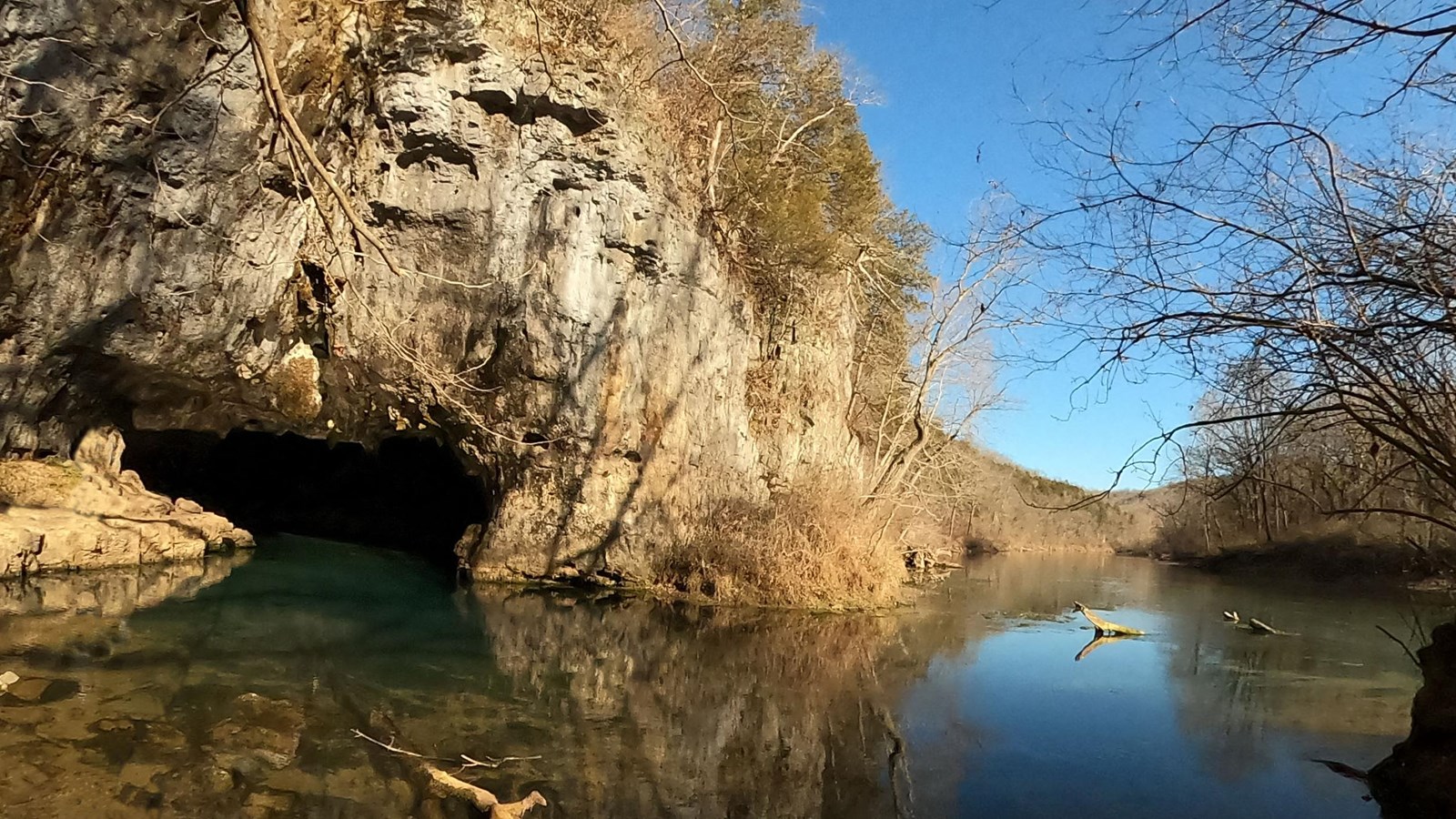 Clear but blueish water with cave opening on left, surrounded by grey rock and a hill
