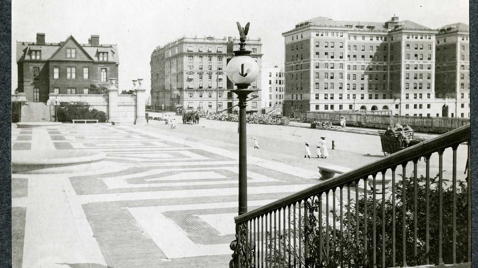 Black and white of stone area with fountains and people walking on it, buildings in distance