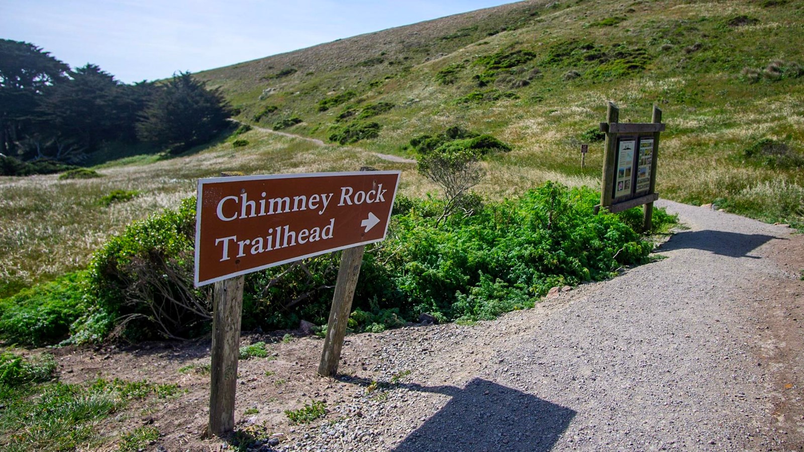 A trailhead sign next to a gravel path that leads past a wooden info kiosk to green, open grassland.