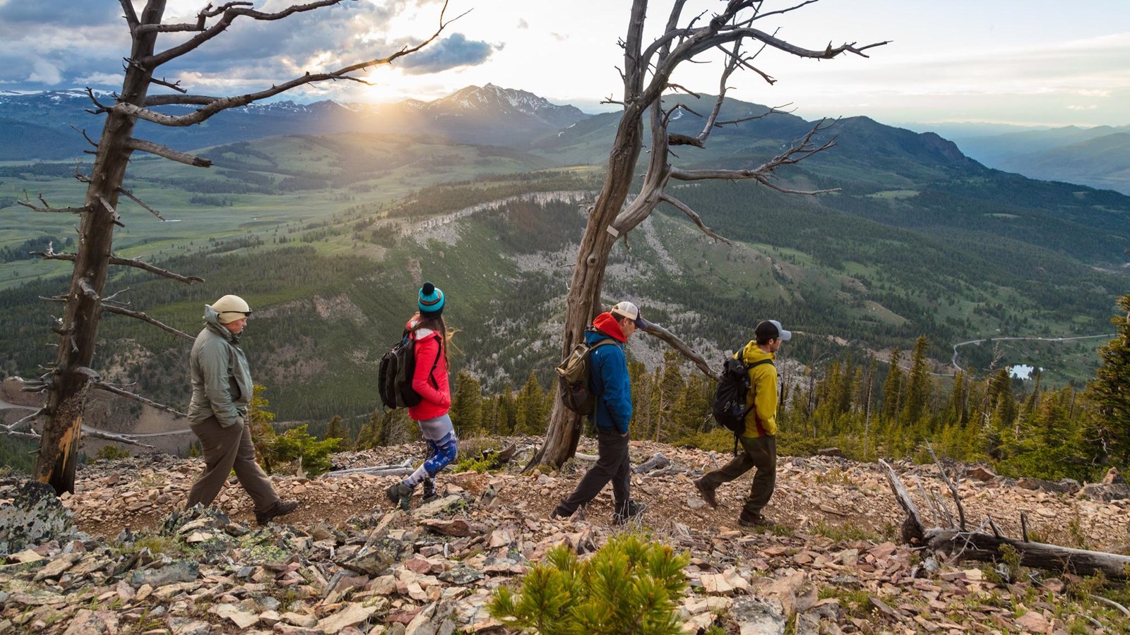 Four people hike across a talus slope with the sun setting behind a mountain in the distance.