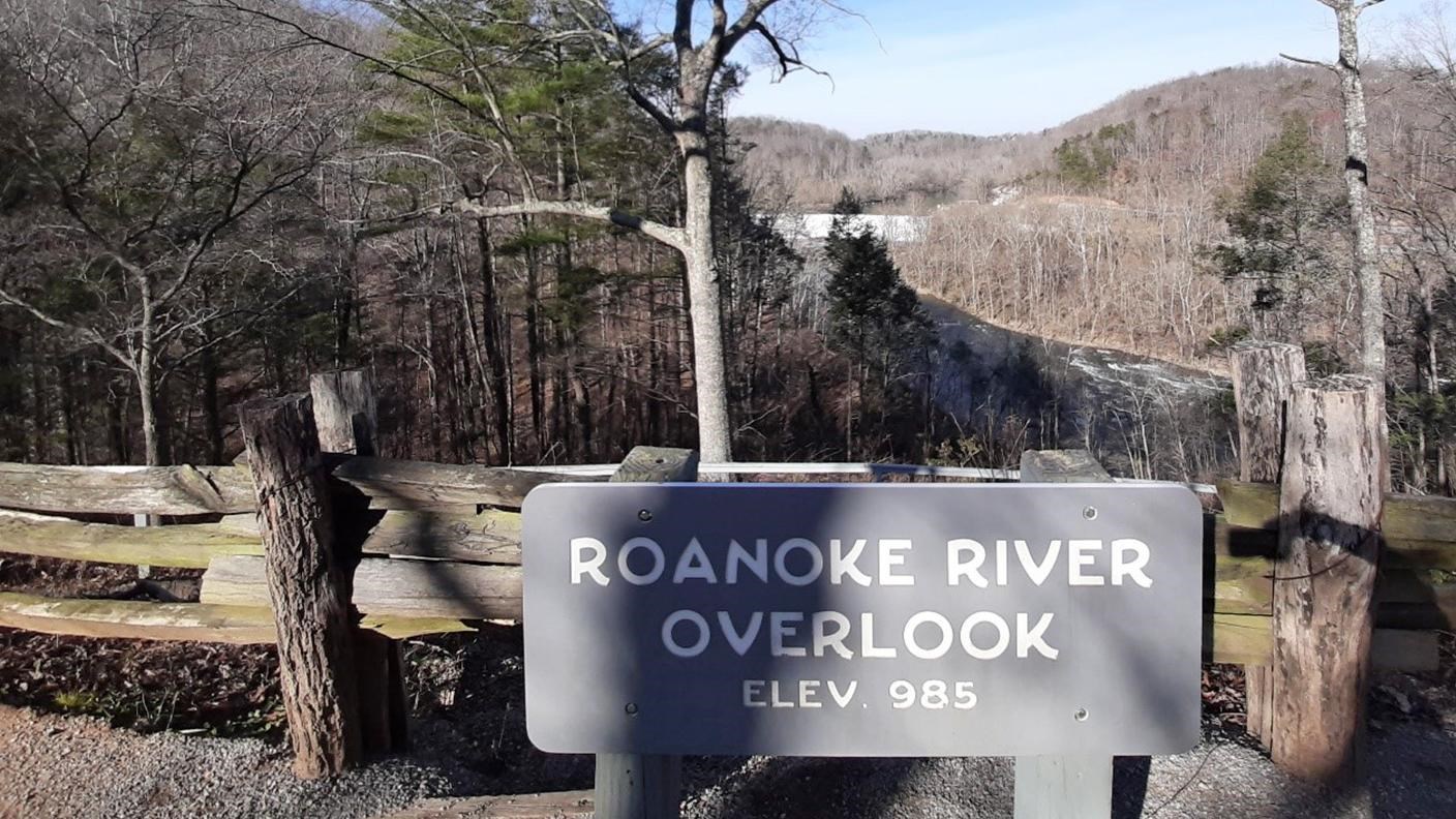 A sign reading Roanoke River Overlook, elevation 985 feet in front of a view of rolling mountains.