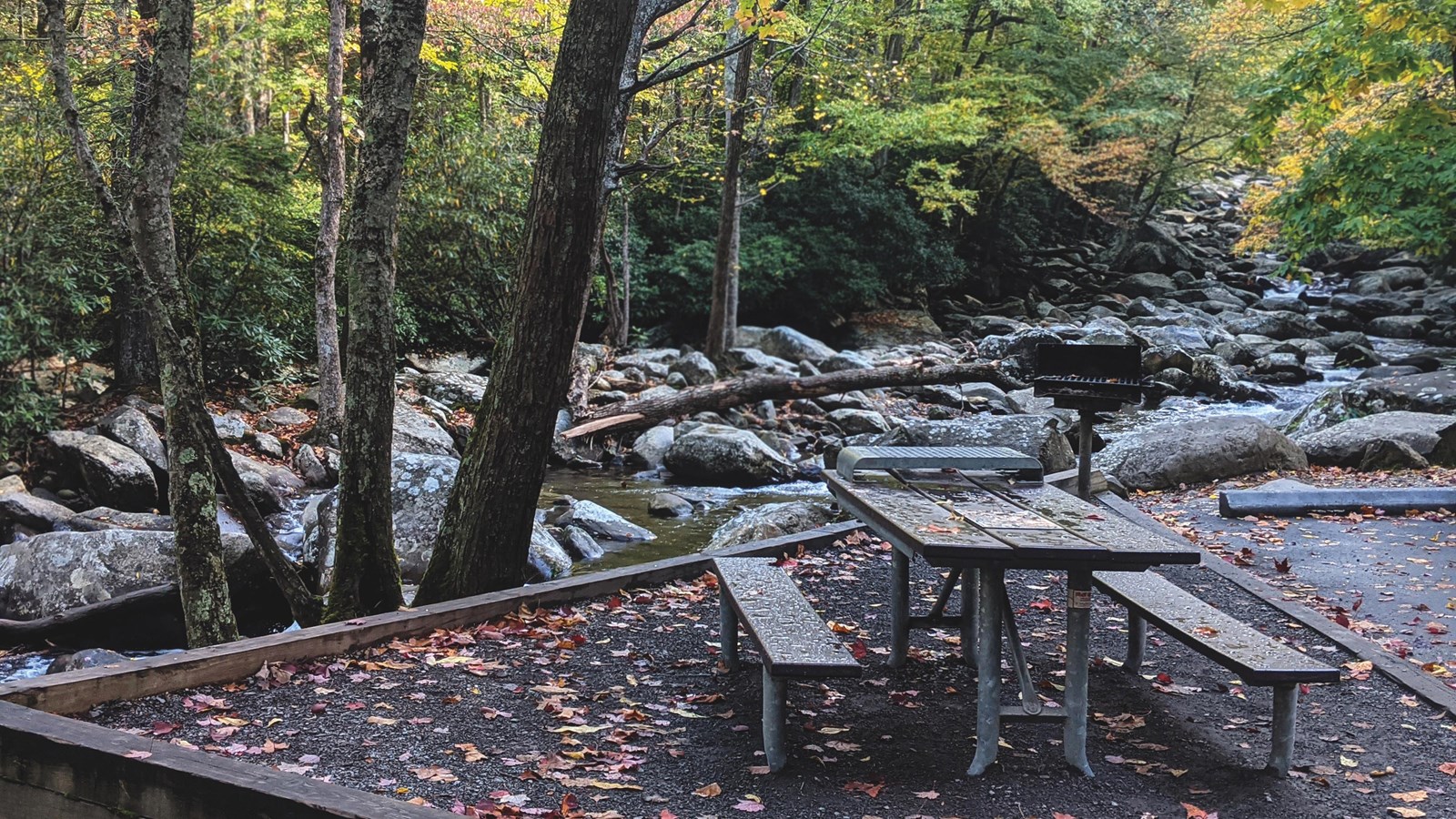 A wooden picnic table on gravel next to a parking space along a rocky river and near trees.