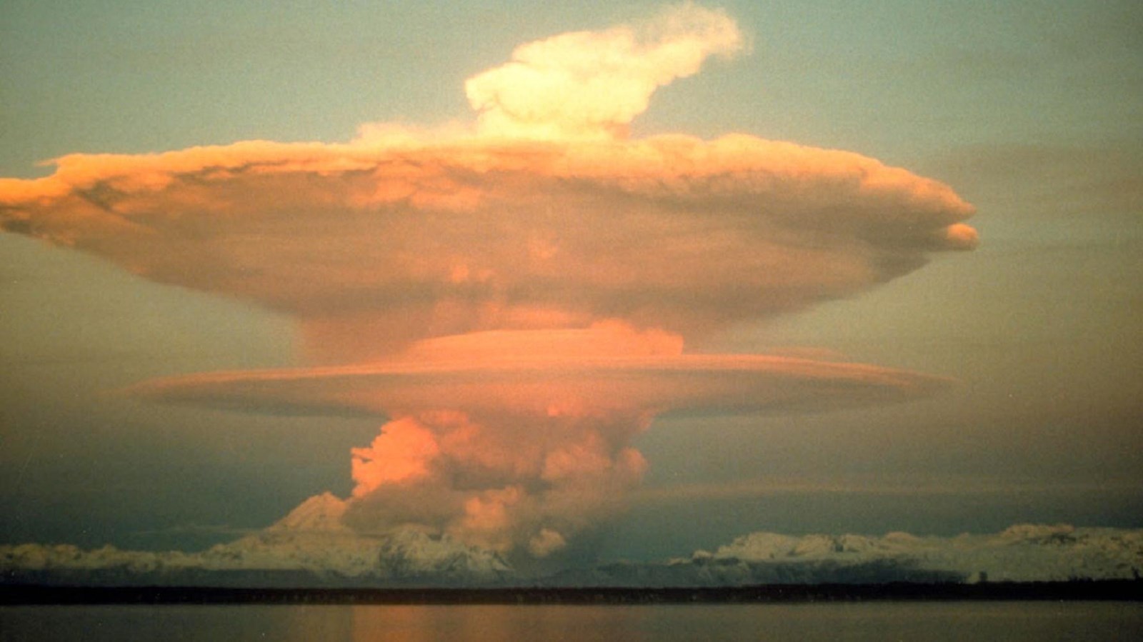 a large ash plume rises from a snow-covered volcano along the ocean