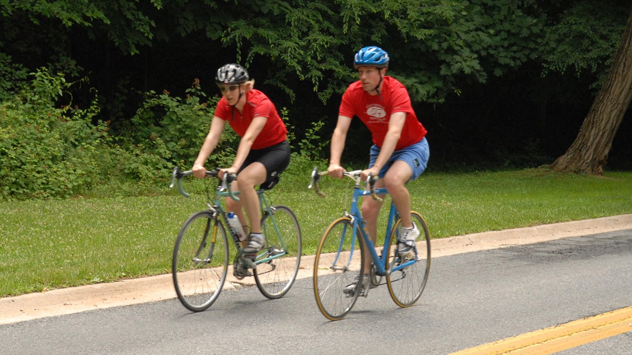 Two bicyclists ride next to each other wearing matching red shirts 