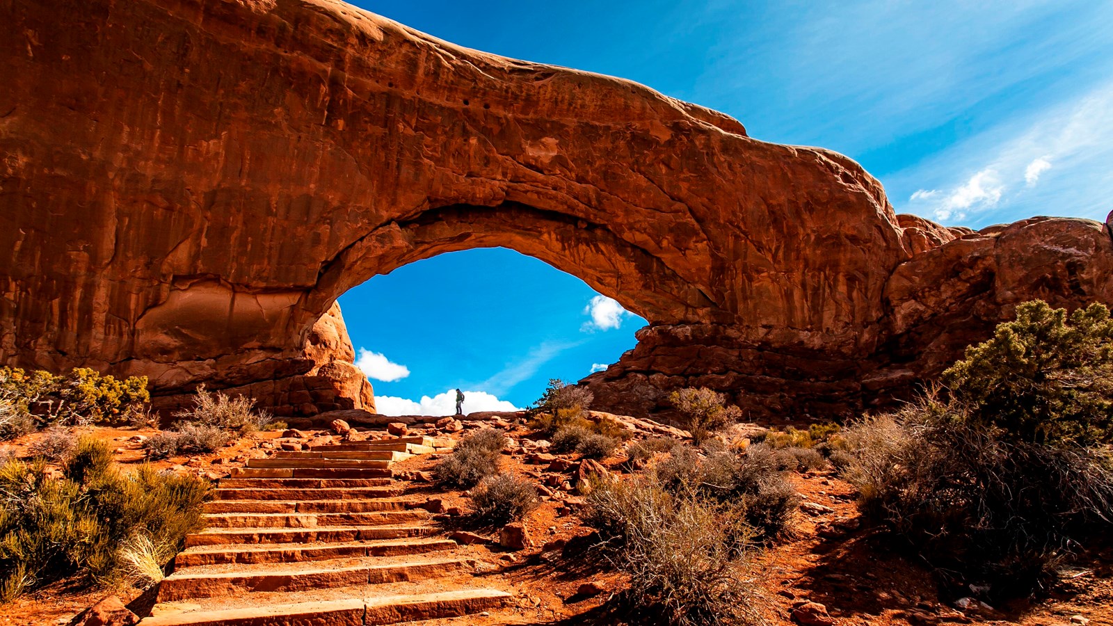 rock steps lead to an orange stone arch towering over a tiny person standing beneath it