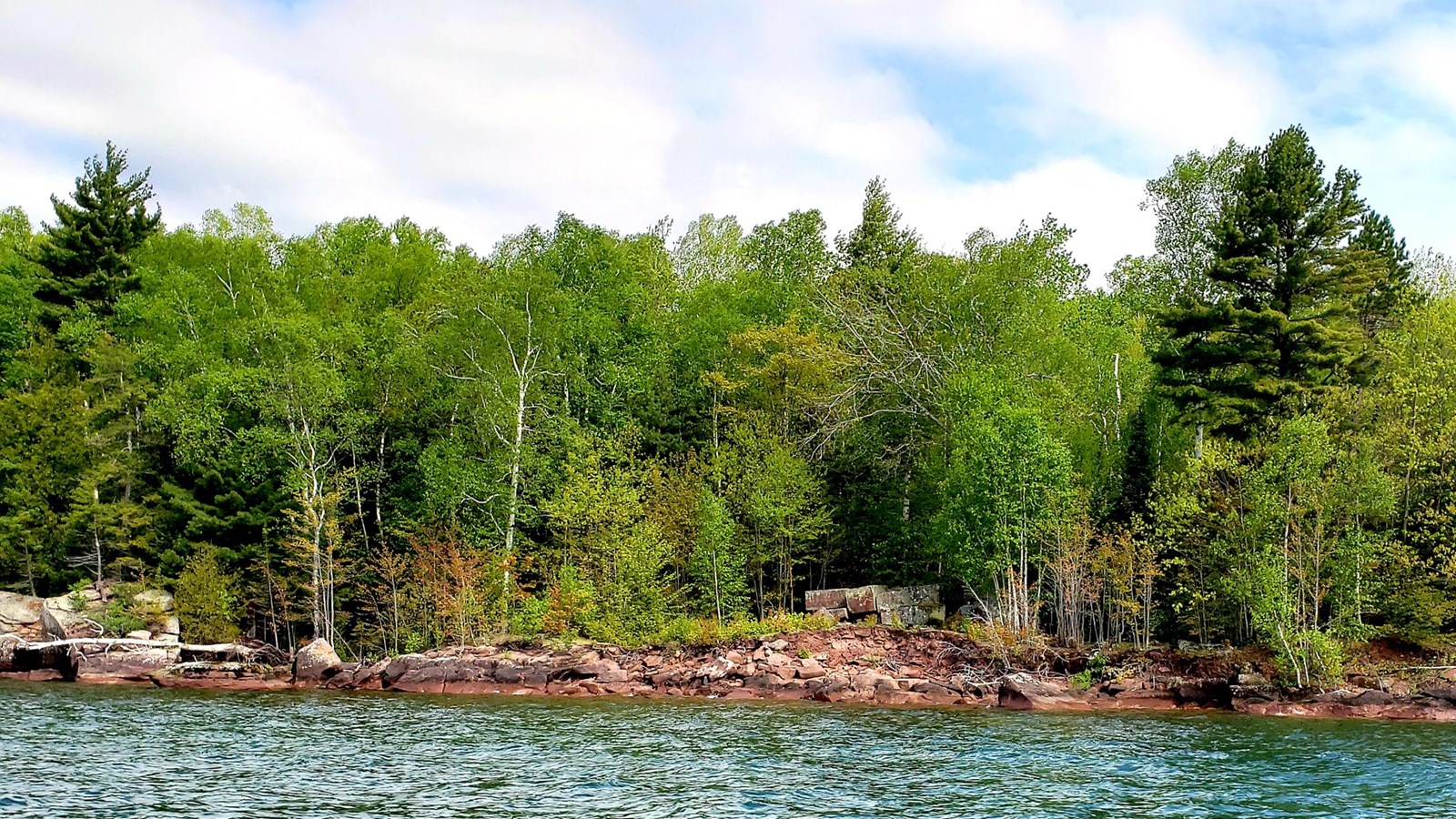 Sandstone shore with trees and blocks, green water in front and cloudy sky.