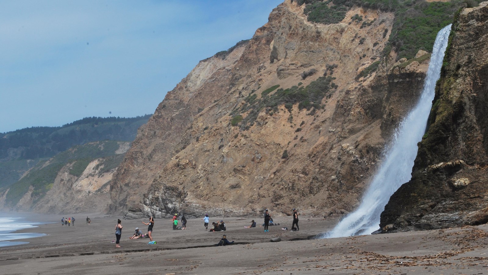 Twenty visitors playing and resting on an ocean beach near the base of a waterfall on the right..