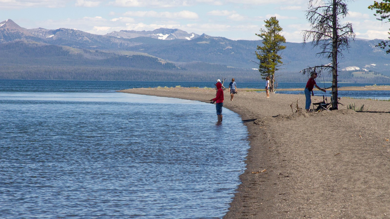 People on a sandy beach of a lakeshore with mountains in the background.