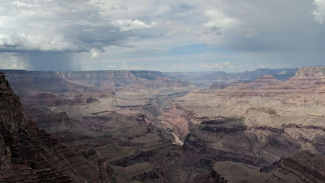 A dramatic canyon vista of cliffs, slopes, and valleys. A storm drops rain in the distance.