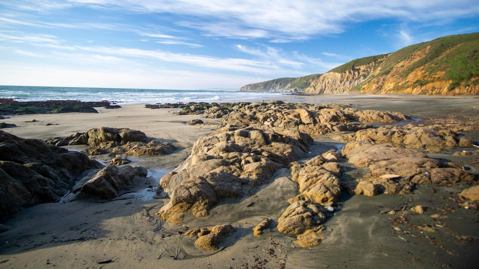 Low-profile rocks rise from a sandy beach. Cliffs reflect afternoon golden light in the background.