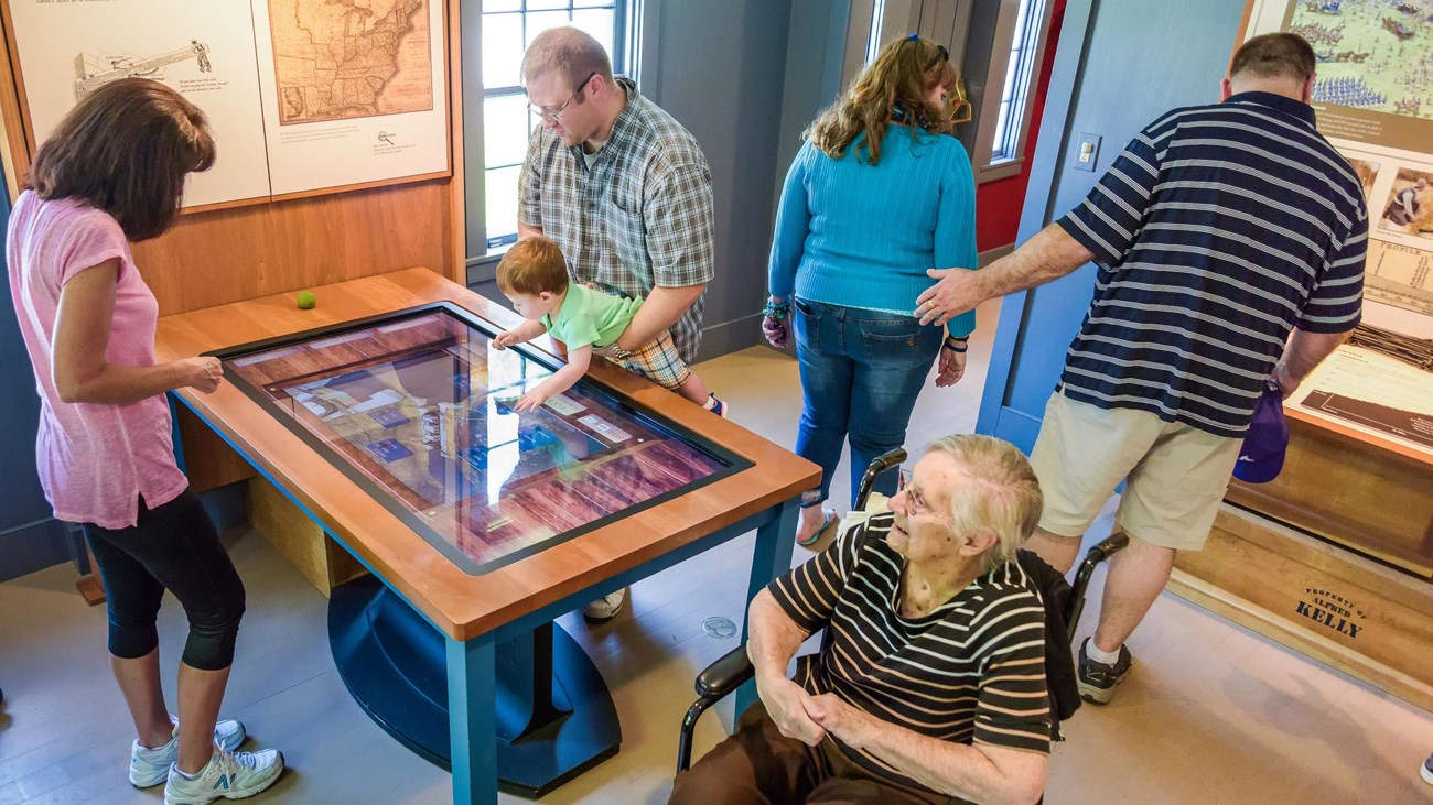 Man holds a child over a touch table as four other adults look at exhibits; one uses a wheelchair.