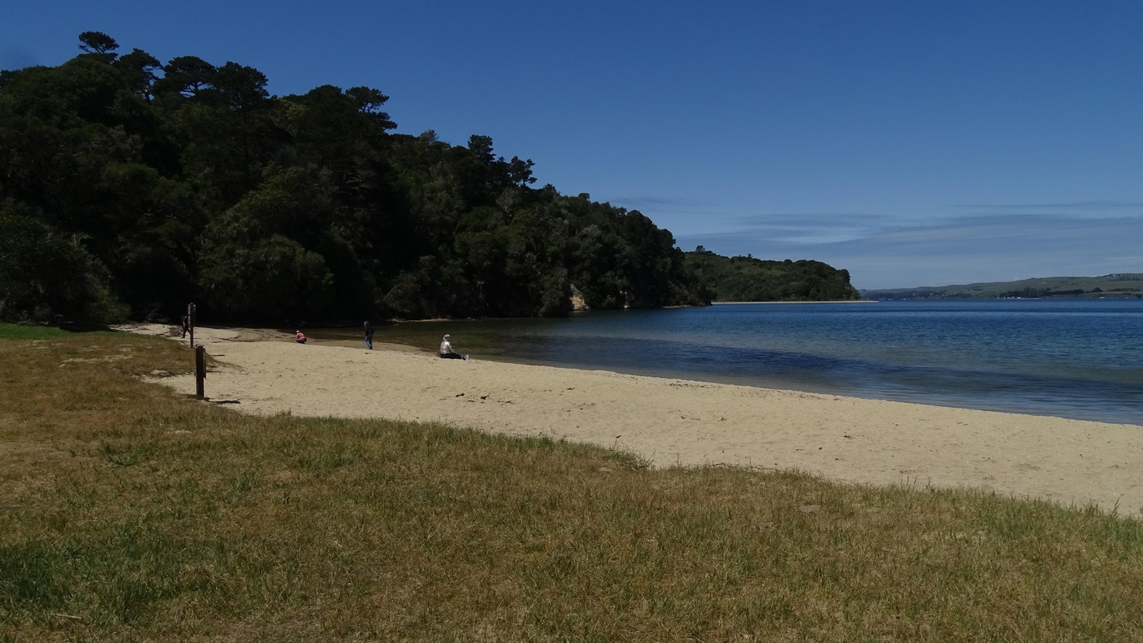 A few visitors sit or walk on a sandy beach adjacent to a blue bay. A forested hill is on the left.